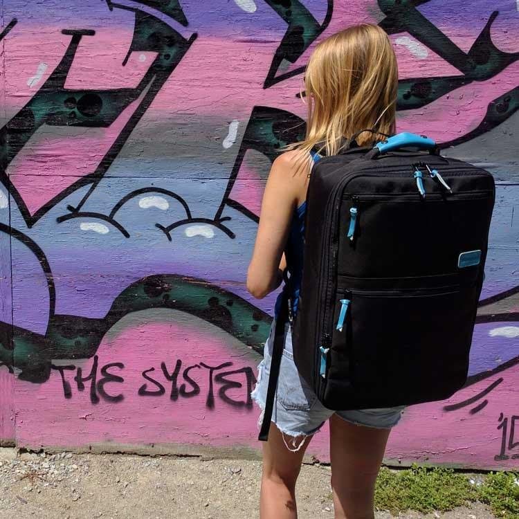 Lady standing with back to front suitcase on shoulders, blonde hair, graffiti wall