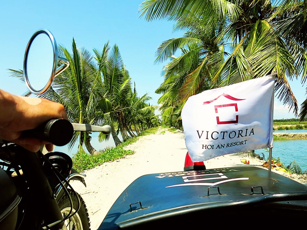 Victoria Hoi An, Vietnam, rural dirt road, side cart motorcycle, palm trees lining the road, water on each side, little flag with resort logo
