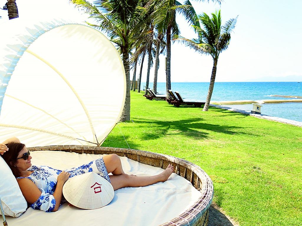 Victoria Hoi An, Vietnam, woman relaxing on the shaded day bed, view of the green grass, water and palm trees