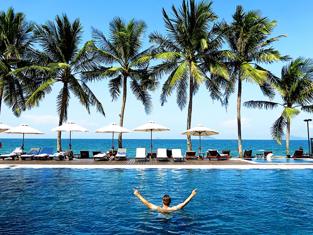 Victoria Hoi An, Vietnam, man with arms up in standing in the pool, view of the sun loungers, umbrellas and palm trees