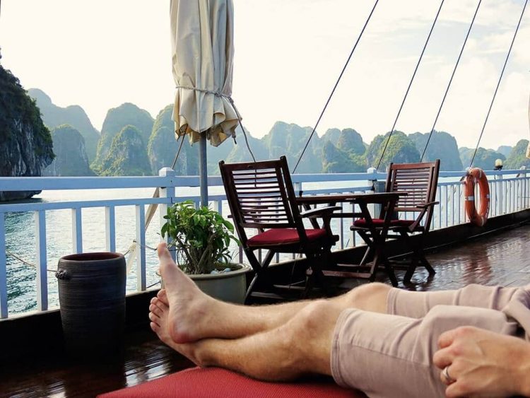 Man sitting laying on the day bed on the deck of the Halong Bay cruise boat, dining set, folded umbrella and view of the bay with rocky islands