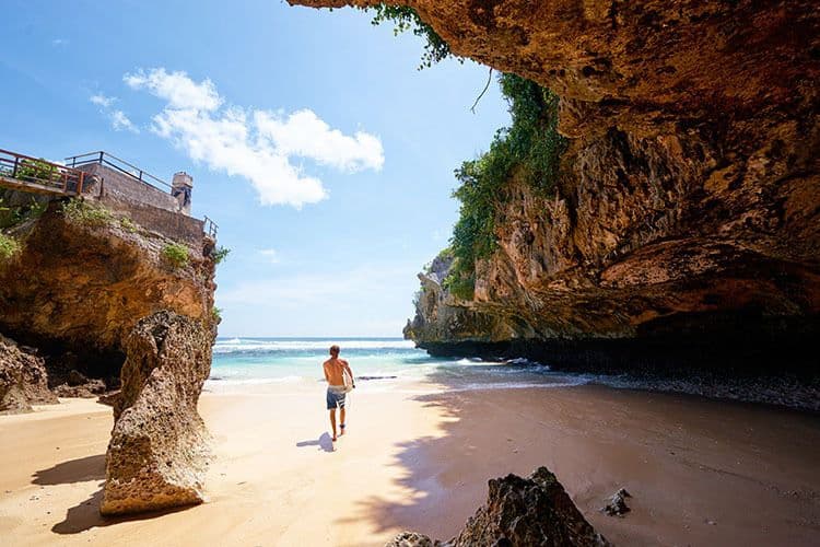 what to do in Southern Bali - Uluwatu Beach, man walking towards the water with a surf board, cave and rocky walls on each side