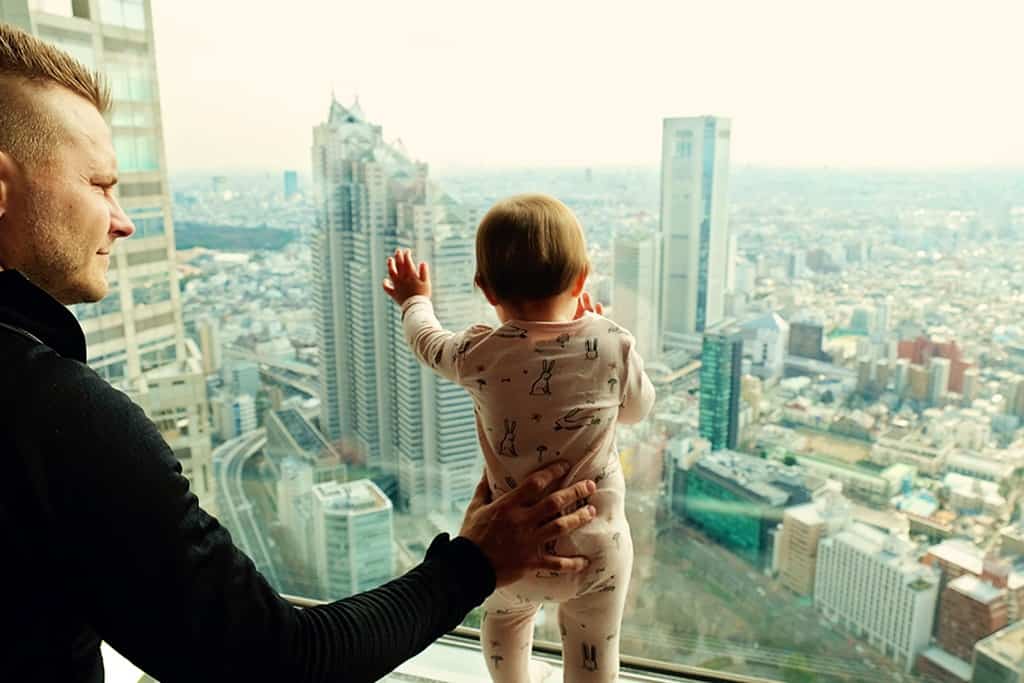 Government Building Tokyo Japan, father holding a baby standing against a window with a view over city
