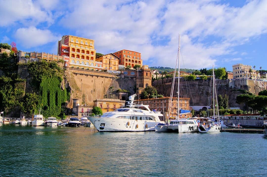 Sorrento Italy, large speed and siling boats parked at the harbour, buildings on the shoreline and high above on the cliff tops