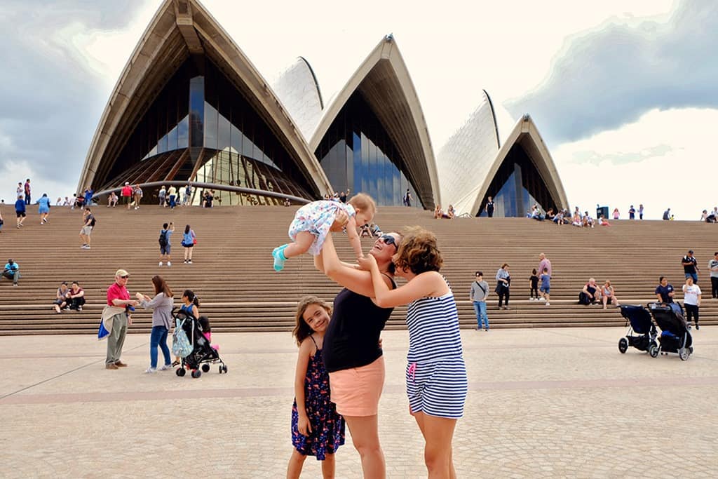 Woman holding a baby in the air smiling, two daughters, people on the steps of the Sydney Opera House, Australia