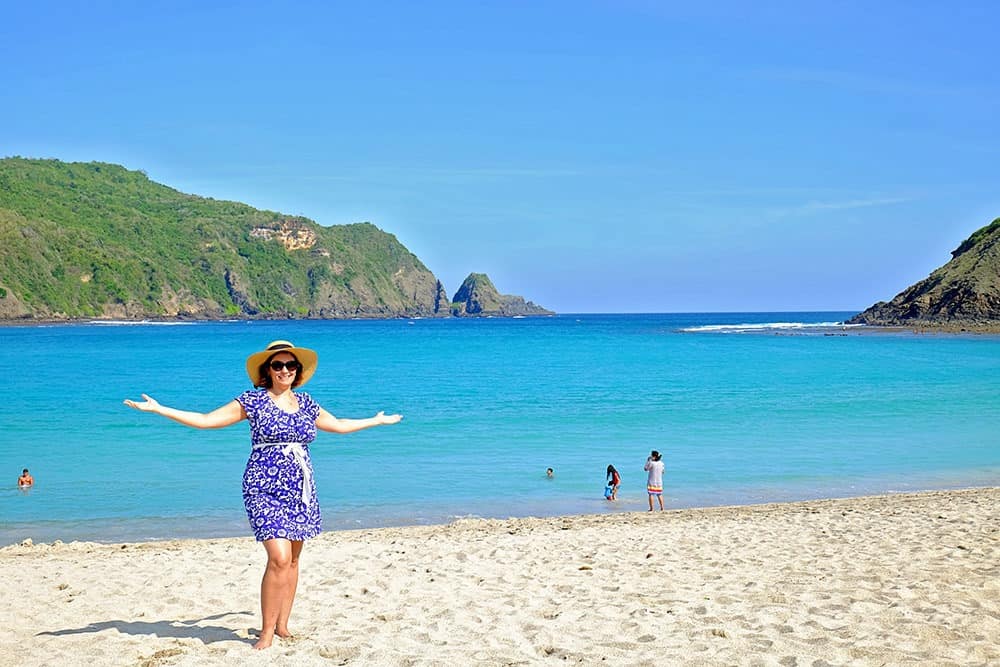 Lombok, Indonesia, woman in blue dress, sunglasses, hat, standing with arms up at the sandy beach, water