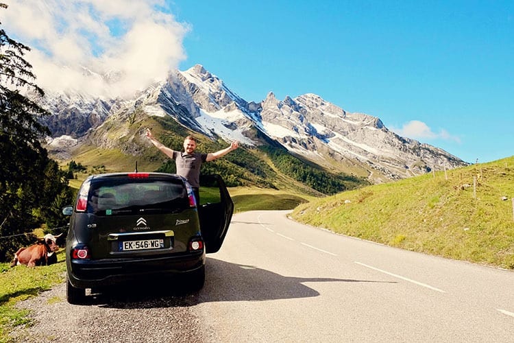 Man standing in the door of the Citroen car, alpine drive in France, cow, mountains with snow covered tops