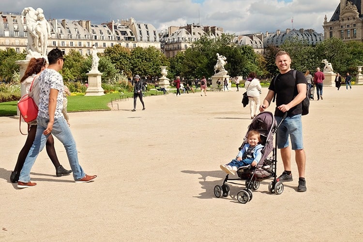 man standing holding a pram, baby in the pram, public park in Paris, France, people walking around