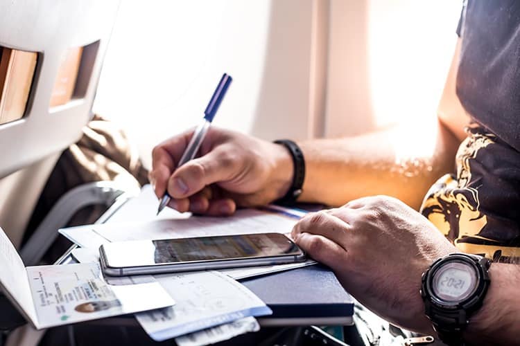 Man sitting in the economy seat in the airplane filling out documents, phone, passport open, pen in hand