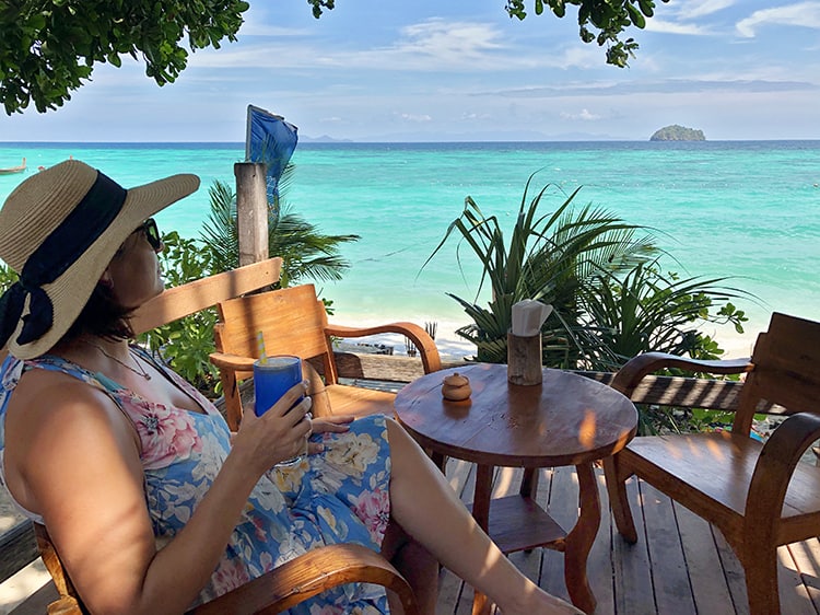 Woman in a hat, blue dress and sunglasses sitting in a restaurant at the beach holding a cocktail and looking out on the light blue water