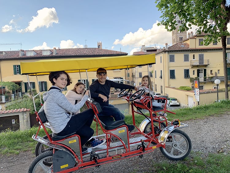 Lucca with Kids, Italy, man and three daughters on a four seater red bicycle with yellow roof, gravel road, buildings in background