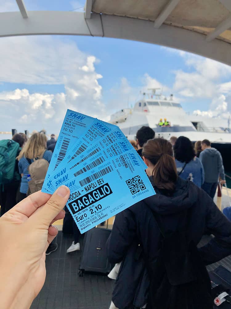 A hand holding blue tickets for the ferry Procida to Napoli, people lining up to the ferry