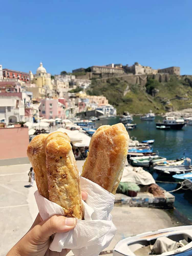 Lingua Procida, Italy, hand holding pastry treats, fishing boats and Marina Corricelli in the background
