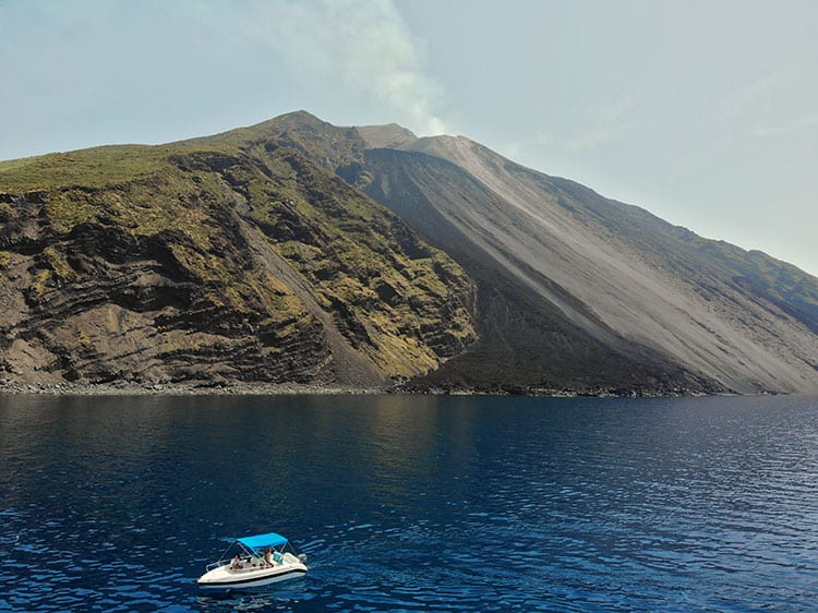 Stromboli Island, Italy, small white boat with blue roof in the water, big volcano island in background