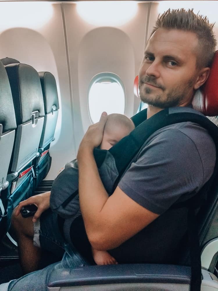 Man sitting in the seat on the airplane holding a baby in the baby carrier