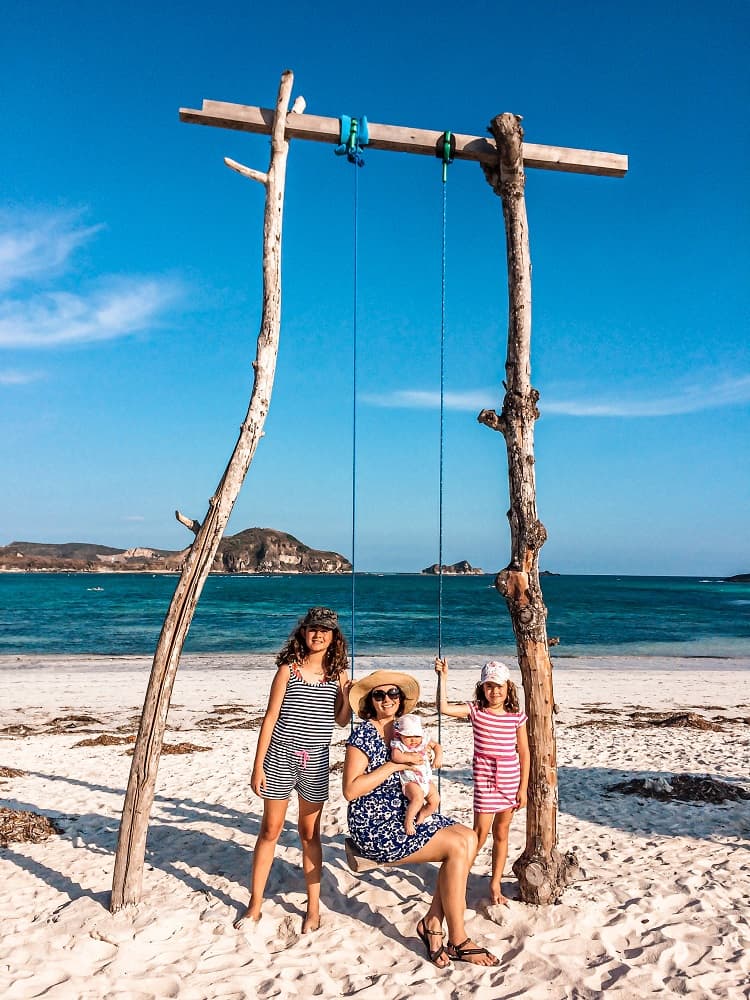 Is it hard to travel with a newborn baby, beach swing, mother sitting holding a baby, two daughters one on each side