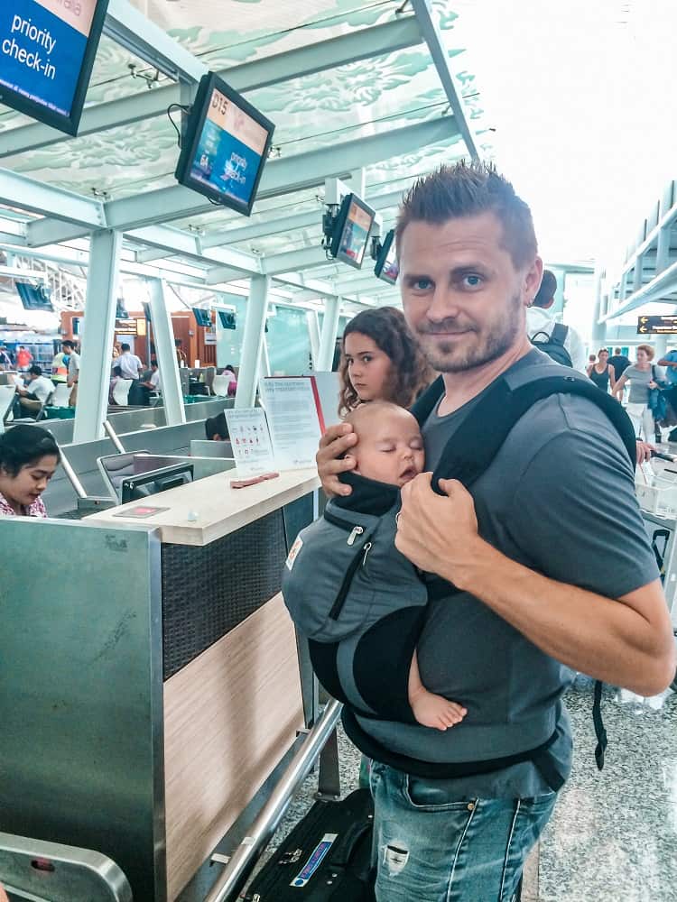 Man holding a baby in the baby carrier at the luggage check in at the airport, man smiling, baby sleeping