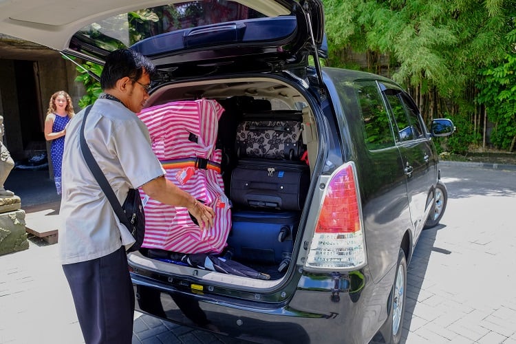Man packing luggage into the back of a black van, teenager waiting at the entrance of the resort