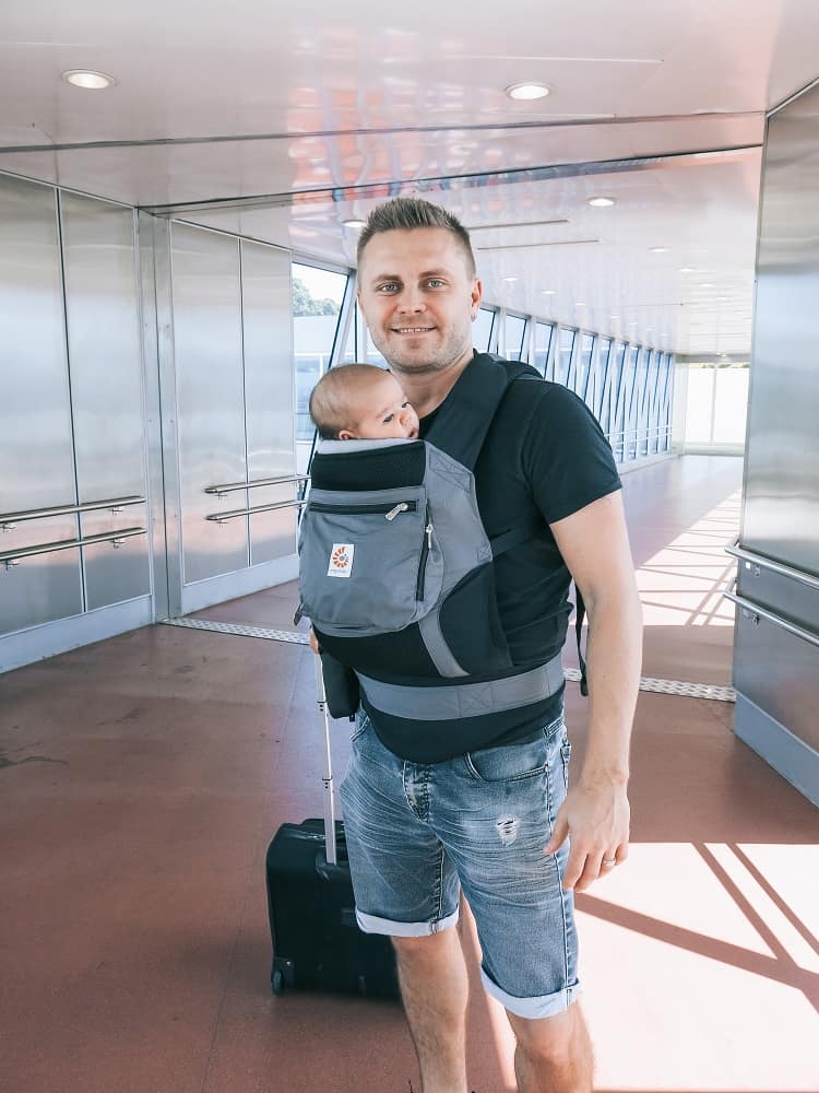 Man standing in the hallway in the airport, holding a baby in the baby carrier.