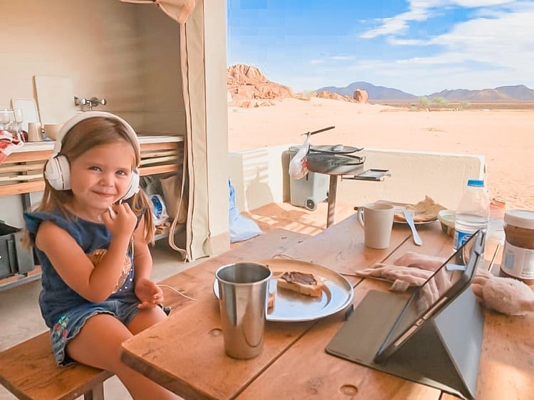 Where to Stay in Sossusvlei with Kids - Dessert Quiver Camp Namibia, young girl eating breakfast, watching iPad, desert in background