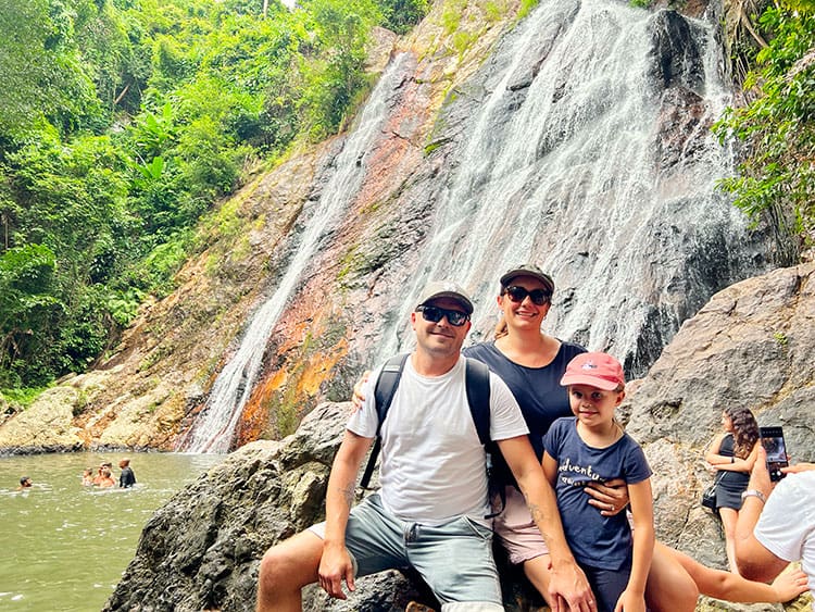 Namuang Waterfall 1, Koh Samui, Thailand, family posing in front of the waterfall, sitting on the rocks, some people swimming bellow the waterfall