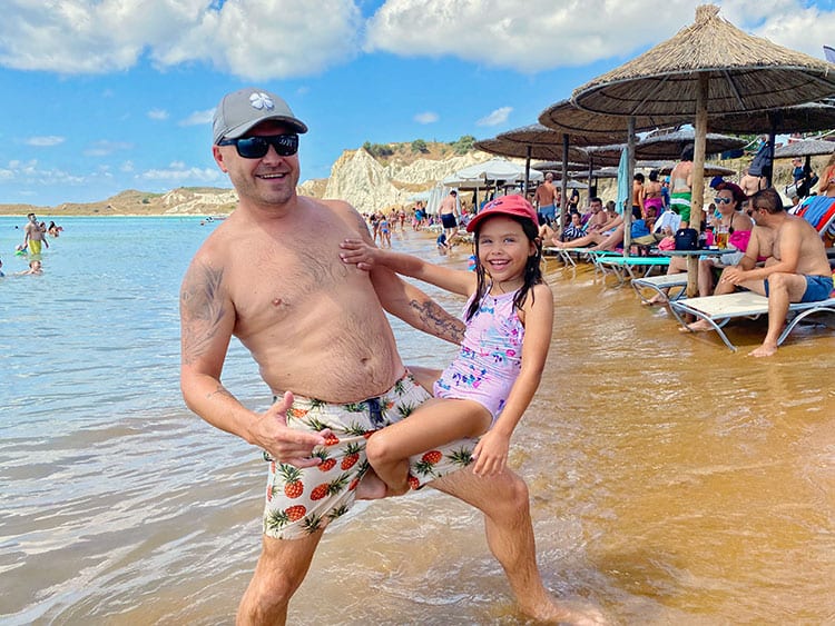 Xi-Beach-Kefalonia, Things to do in Kefalonia, Greece, father and daughter standing in the water, tourists, sun loungers and beach umbrellas 