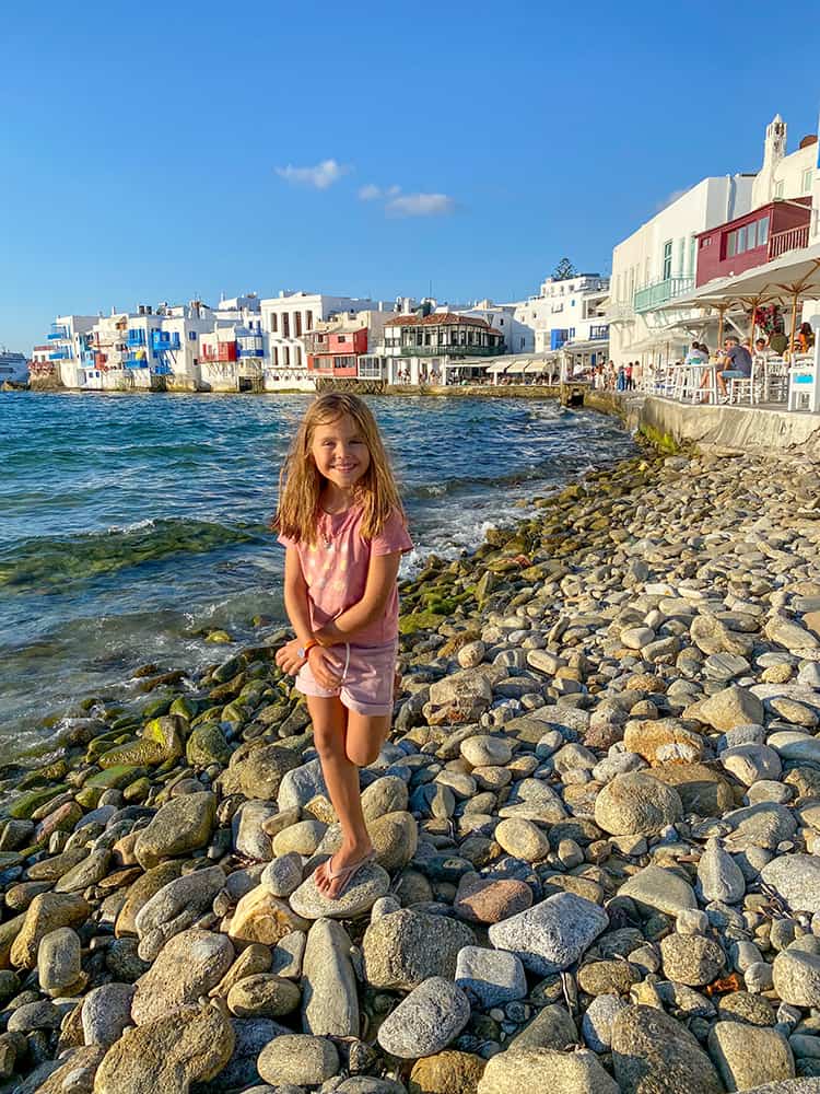 Mykonos in September, Greece - Little Venice, young girl on the rocky beach, Little Venice in the background