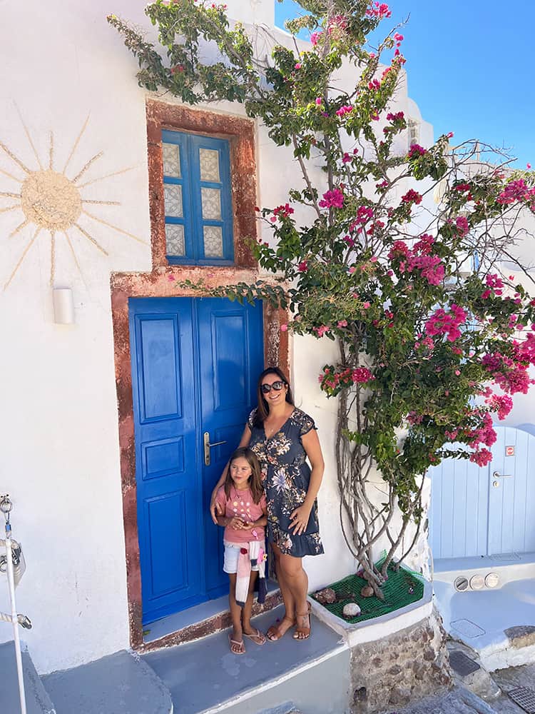 Santorini in September, Greece - Mother and daughter posing in front of a white building with blue door, pink flower tree