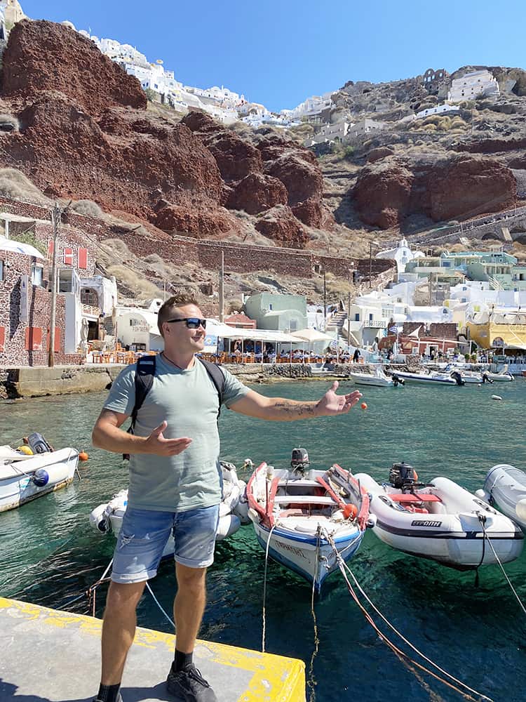 Santorini in September, Greece - man posing pointing to the boats in the harbour, Oia town up the top of the mountain