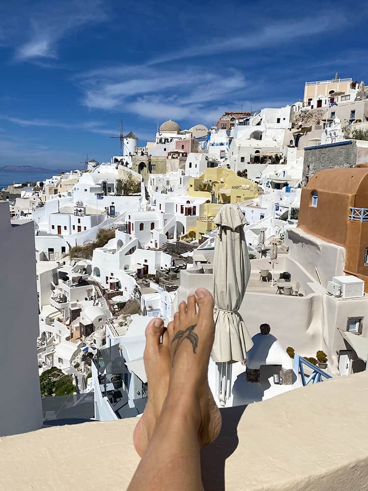 Santorini in September, Greece - womans legs pointing towards a view of the buildings on the side of the hill